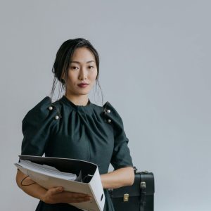 Woman in a dark dress holds a Human Resource Management binder beside a suitcase near a gray wall.