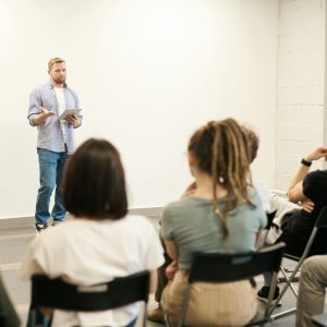 Man presenting "Human Resource Management" to seated audience in a bright, white-walled conference room.