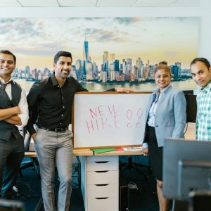 Four colleagues stand by a whiteboard labeled "NEW HIRE!!" in an office with a city skyline view.