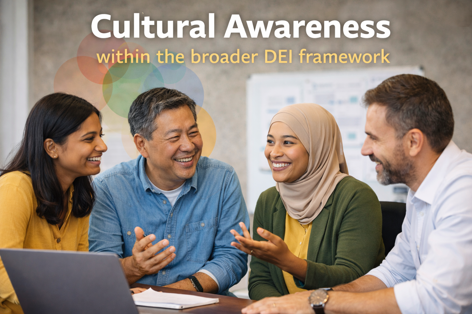 Four colleagues sit at a table, smiling and talking beneath a sign reading "Cultural Awareness in DEI.