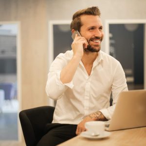 Smiling man in white shirt at desk with laptop, talking on phone.