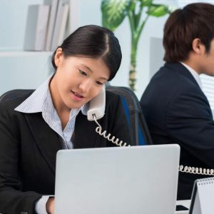 Businesswoman on phone at laptop; man beside her focused on his desk, both working in office.