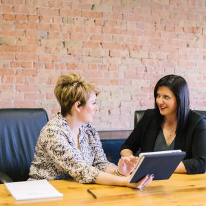 Two women talking at a desk in a modern brick office; visible text: Coaching and Mentoring - Self Study.