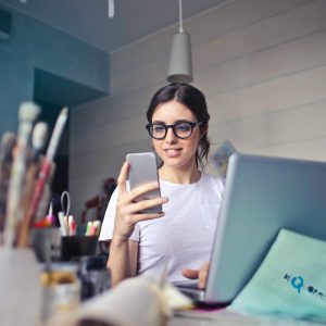 Woman with glasses studies “Communication Strategies” on her phone at a desk with art supplies.