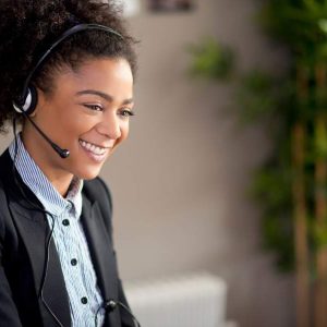 Smiling woman in headset at desk with plant, using Customer Service - Self Study program.