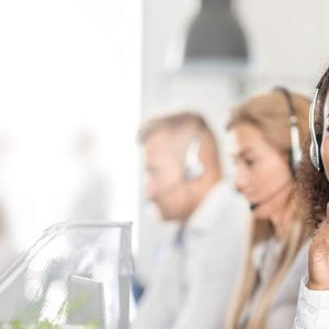 Woman wearing headset studies “Customer Support - Self Study” book; coworkers visible in background.