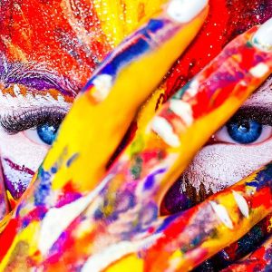 Blue-eyed person with painted face hides behind book titled “Developing Creativity - Self Study.”.