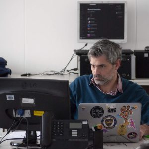 Gray-haired man studies “Facilitation Skills - Self Study” course at desk with monitors and laptop.
