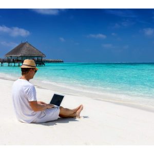 Person on sandy beach using a laptop, clear blue water and huts visible in the background.