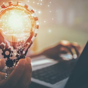 Hand holding a glowing brain-shaped bulb beside laptop, with text: Marketing Basics - Self Study.