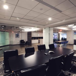 Conference room with long table, black chairs, and glass walls overlooking office workspace.